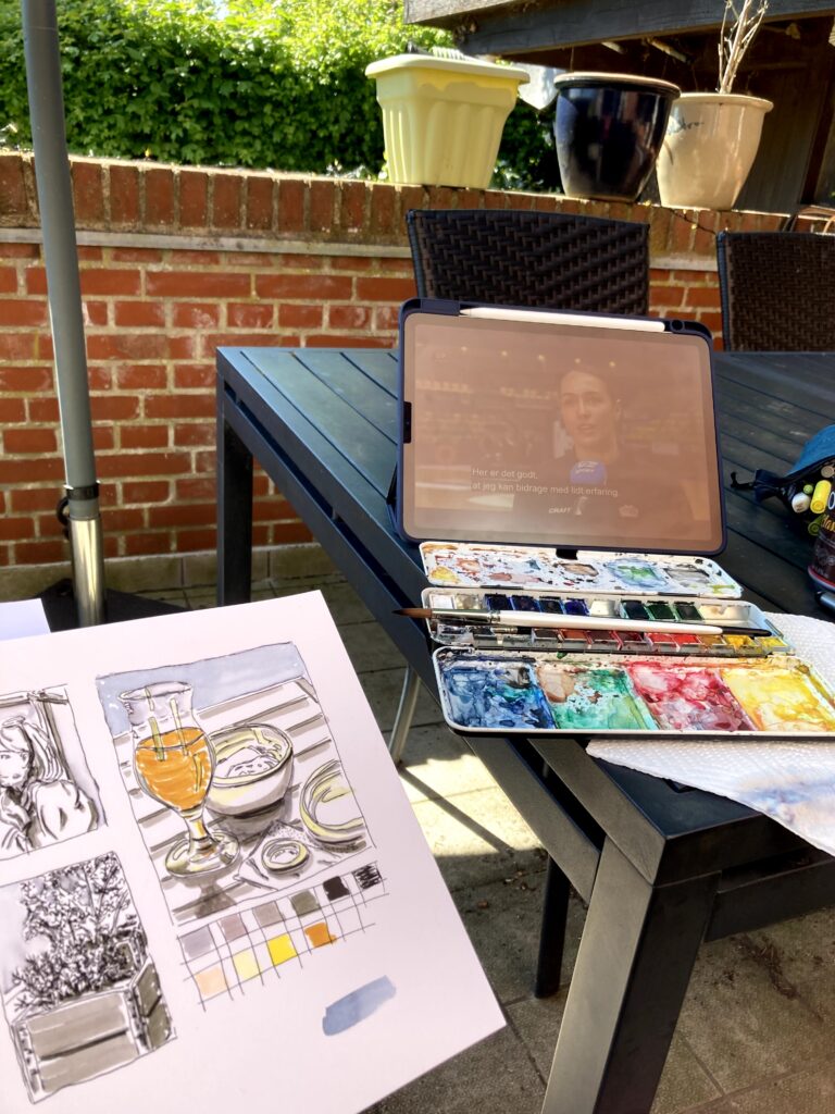 A water colour palette on a garden table and a sketch next to it telling the story of a person sitting outside on a terrasse and drawing
